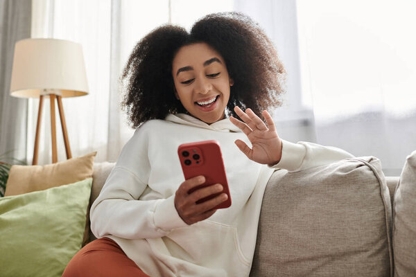 A young woman in a cozy outfit happily chats on her smartphone while relaxing in her living room.