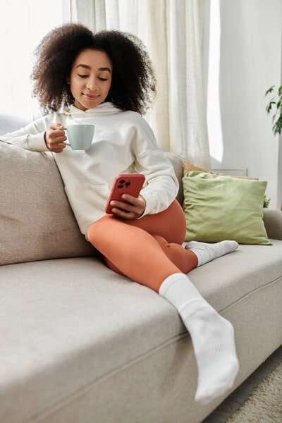 Молодая женщина расслабляется на диване, потягивая чай и просматривая свой телефон в уютном одеянии.