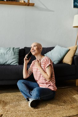 Alopecia hastası neşeli genç bir kadın yerde oturuyor, telefonla konuşurken gülümsüyor..