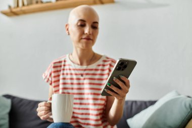 Kel kafalı genç bir kadın çayını yudumluyor, akıllı telefonuyla rahat bir yere dalmış..