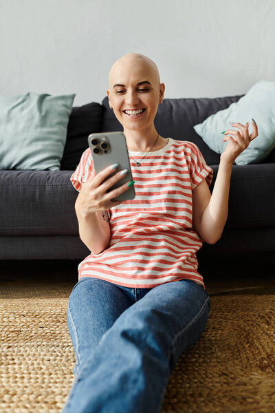 Молодая лысая женщина с сияющей улыбкой наслаждается своим смартфоном на мягком коврике дома.