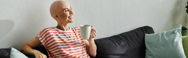 A happy bald woman enjoys her coffee while relaxing on a comfortable sofa filled with cushions.
