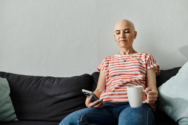 Молодая женщина с алопецией расслабляется на диване, потягивая чай, проверяя телефон..