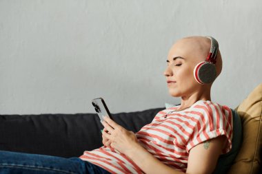 Kel bir kadın kulaklıkla müzik dinlemekten hoşlanır, rahat bir kanepede telefonuna dalmıştır..