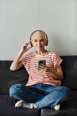 A joyful bald woman with alopecia relaxes on a couch, immersed in her music. clipart