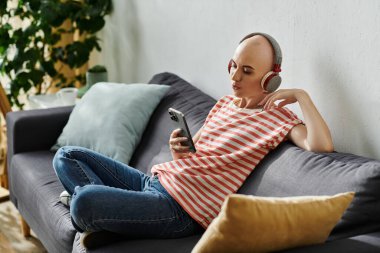 A beautiful bald woman listens to music while lounging on a comfortable couch at home. clipart