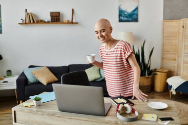 Alopecia hastası neşeli genç bir kadın laptopuyla temas kurarken kahveyi yudumluyor ve sıcaklığını yayıyor..