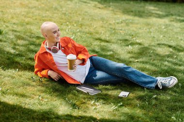 Genç, kel bir kadın, gün ışığında sıcak bir içecek yudumlarken gülümseyerek çimenlere yaslanıyor..