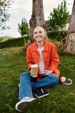 A young bald woman sits on the grass, holding coffee and a notebook, radiating happiness in nature. clipart