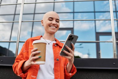 Genç, güzel, kel bir kadın elinde kahve fincanı tutarken telefonuna bakarken gülümsüyor..