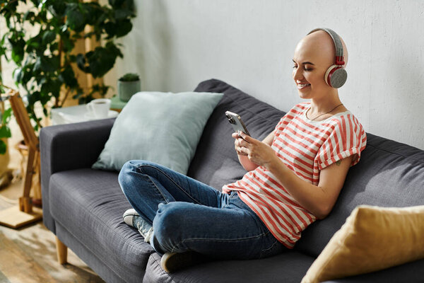 Лысая женщина слушает музыку на телефоне, удобно сидя на удобном диване.