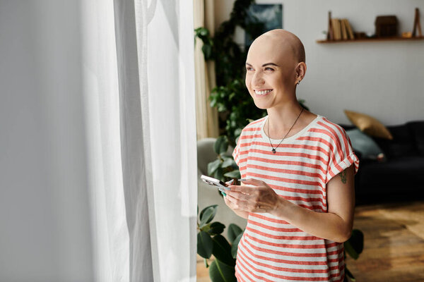 Красивая лысая женщина стоит у окна, радостно общаясь со своим телефоном в ярком пространстве.