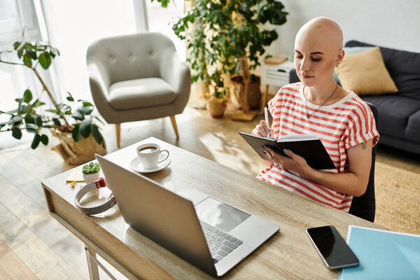 Молодая лысая женщина сосредотачивается на том, чтобы писать в блокноте, сидя за хорошо освещенным столом.