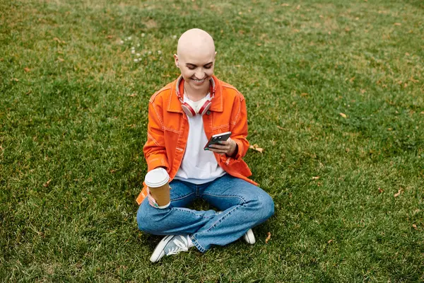 stock image A young woman with a beautiful bald head smiles as she relaxes on the grass, holding coffee.