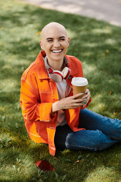 Молодая лысая женщина с сияющей улыбкой пьет кофе, сидя на зеленой траве.
