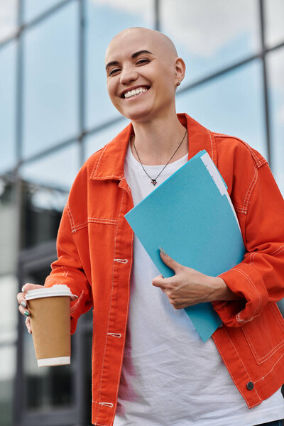 Лысая женщина излучает радость, сжимая напиток и яркие папки под чистым небом.