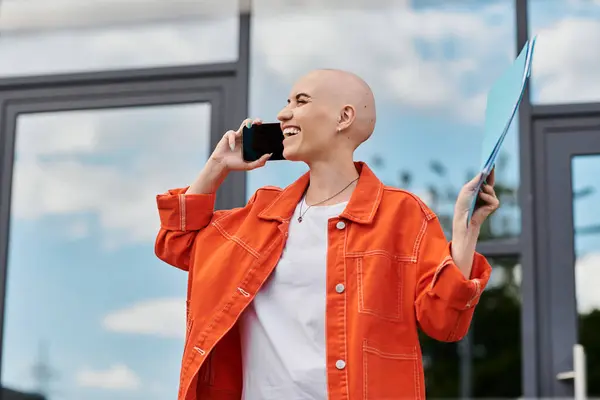 Stock image A confident woman engages in a lively phone conversation while holding documents outdoors.