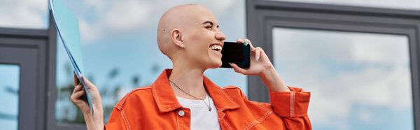 A bald woman joyfully chats on her phone, holding a folder in a vibrant orange jacket.