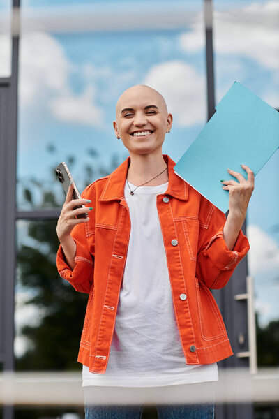 Радостная лысая женщина держит в руках бумагу и телефон, излучая уверенность современной стеклянной структурой.