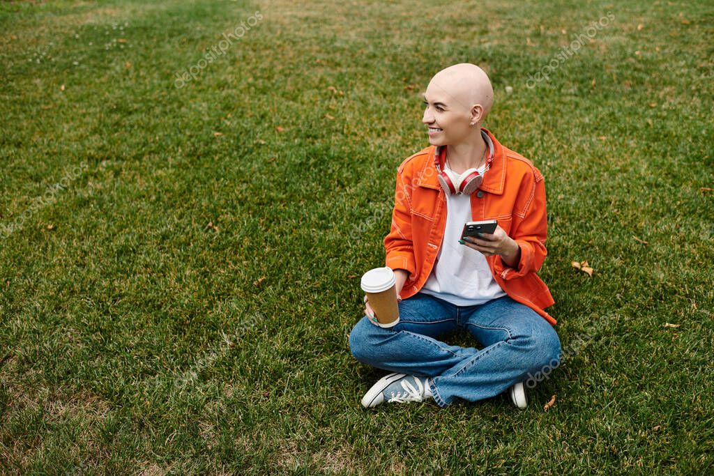 A young woman with a beautiful bald head sits cross legged on the grass, smiling.