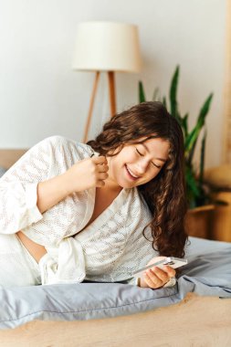 A young beautiful plus size woman laughs while lounging at home, engaging with her smartphone. clipart