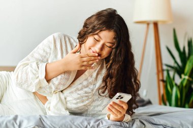 A young plus size woman relaxes at home, engaged with her phone on a cozy bed. clipart