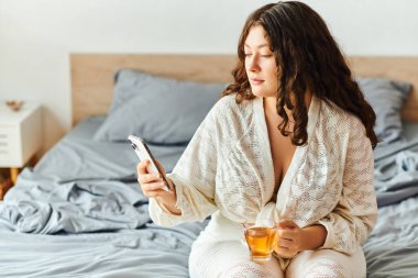Yatakta dinlenen genç bir kadın, rahat ve huzurlu bir ortamda telefonunu kontrol ederken çayını yudumluyor..