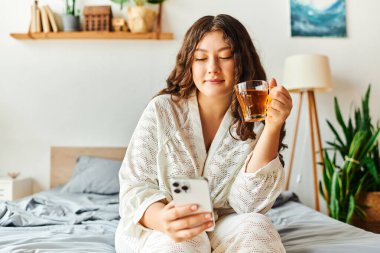 Genç ve iri yarı bir kadın rahat bir odada telefonuyla dolaşırken sıcak bir içkiden zevk alır..