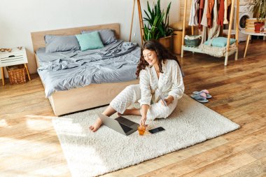 Genç ve iri yarı bir kadın evdeki laptopuyla çalışırken yumuşak halıda dinleniyor..