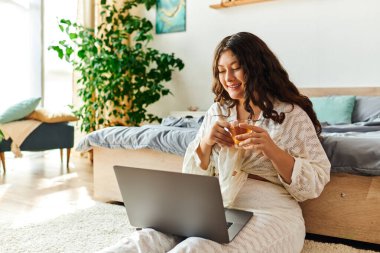 A joyful plus size woman sips tea and smiles while engaged with her laptop in a cozy setting. clipart