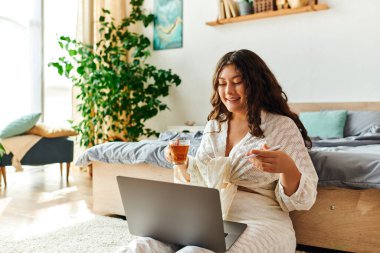 Büyük beden bir kadın evinde rahatlar, çayını yudumlar ve sıcak bir atmosferde laptopuyla meşgul olur..