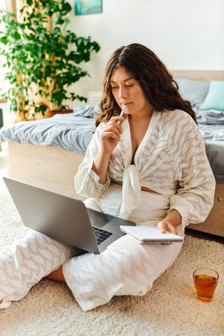 A young beautiful plus size woman thoughtfully writes in a notebook while using her laptop at home. clipart