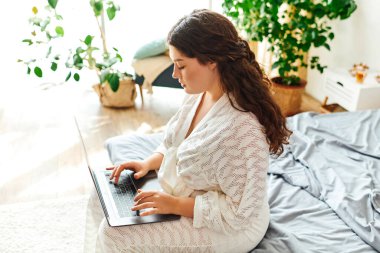 Geniş beden bir kadın, yeşil bir yatakta rahatça otururken dizüstü bilgisayarıyla meşgul olur..