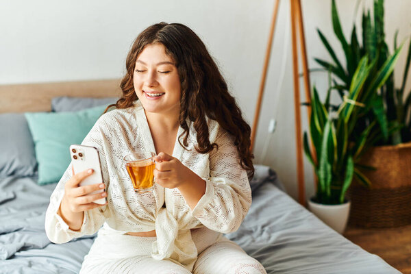 Веселая молодая женщина расслабляется дома, потягивая чай, общаясь со своим телефоном.