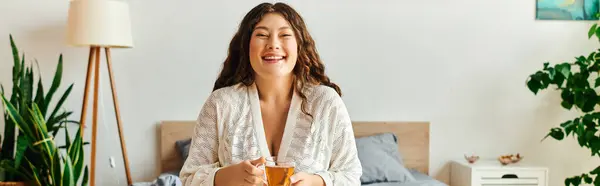 stock image A young beautiful plus size woman sits comfortably, smiling and sipping tea in her inviting bedroom.