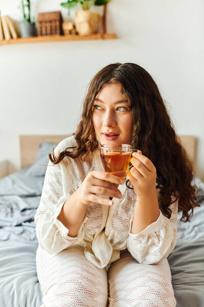 A beautiful young woman sits on her bed, sipping tea and enjoying a peaceful moment at home.