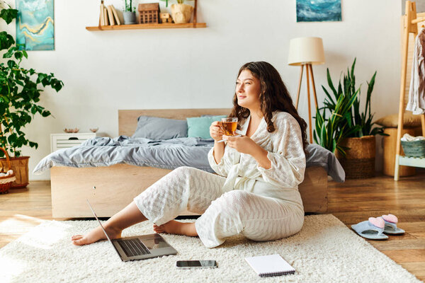 A young plus size woman relaxes at home, sipping tea and working on her laptop.