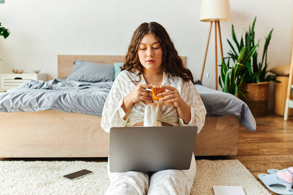 Молодая женщина расслабляется дома, потягивает напиток и фокусируется на своем ноутбуке в уютном месте.