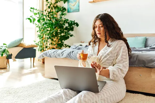 stock image A young plus size woman relaxes at home, sipping tea while focused on her laptop screen.