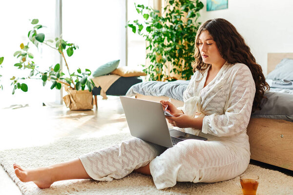 Молодая красивая женщина plus size удобно сидит на полу со своим ноутбуком, занятая работой.