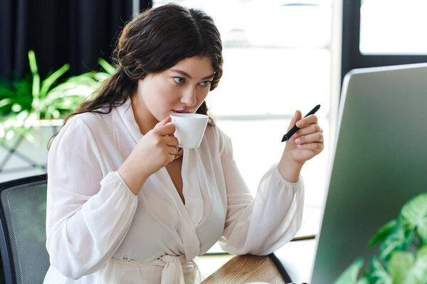 A young beautiful woman savors her tea while focused on her computer work at office.