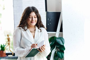 A young plus size woman stands at office, writing in her notebook with a cheerful smile. clipart