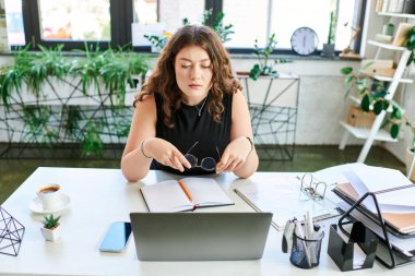 A young businesswoman with curly hair focuses intently on her laptop while organizing notes at her desk. clipart