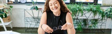 Uzun kıvırcık saçlı neşeli ve iri yarı bir kadın, işyerindeki yemyeşil çiçeklerle çevrelenmiş vazifeleriyle meşgul olur..