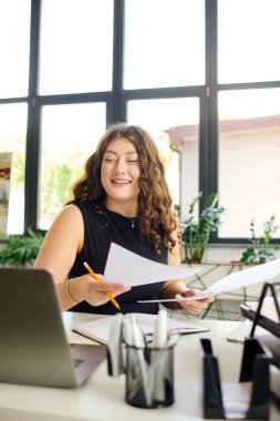 A radiant plus-size woman with curly hair engages in her work, sorting through documents with enthusiasm in a sunlit office. clipart