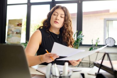 Genç ve cüsseli bir profesyonel masasında oturmuş belgeleri inceliyor ve ilham verici bir ofis ortamının ortasında işe dalmış..