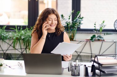 A confident plus-size woman with long curly hair smiles while reviewing documents at her office desk. clipart