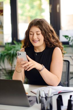 Kendine güvenen kıvırcık saçlı genç bir kadın modern bir ofis masasında otururken akıllı telefonuna odaklanıyor..