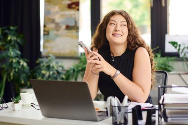 Kendine güveni yüksek kıvırcık saçlı bir kadın modern bir ofis alanında akıllı telefonuyla meşgul oluyor..