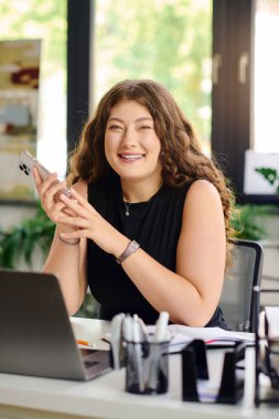 Uzun kıvırcık saçlı neşeli ve iri yarı bir kadın akıllı telefonunu parlak bir ofis alanında kullanıyor. Kendine güveni ve yaratıcılığı somutlaştırıyor..
